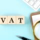 Wooden blocks spelling VAT next to a magnifying glass, pen, and financial spreadsheet on a blue background.