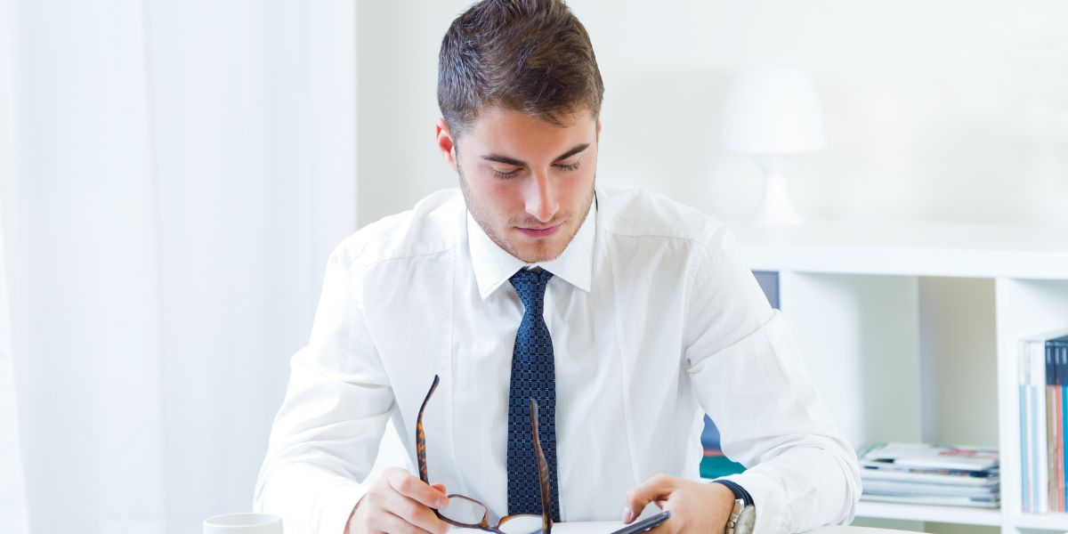 A businessman in a white shirt and tie looking down while holding eyeglasses at his office desk.