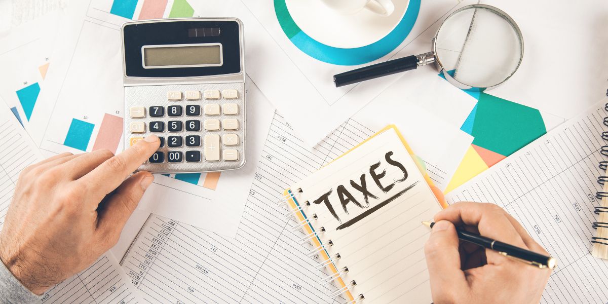 Top view of hands using a calculator and writing "TAXES" in a notepad next to financial charts.